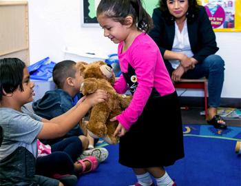 Elementary-aged girl hands a teddy bear to a classmate while a female teacher watches