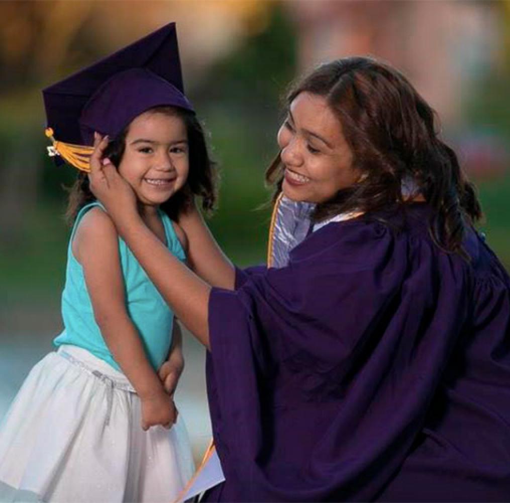 Woman in a graduation gown crouching next to a girl with a graduation cap