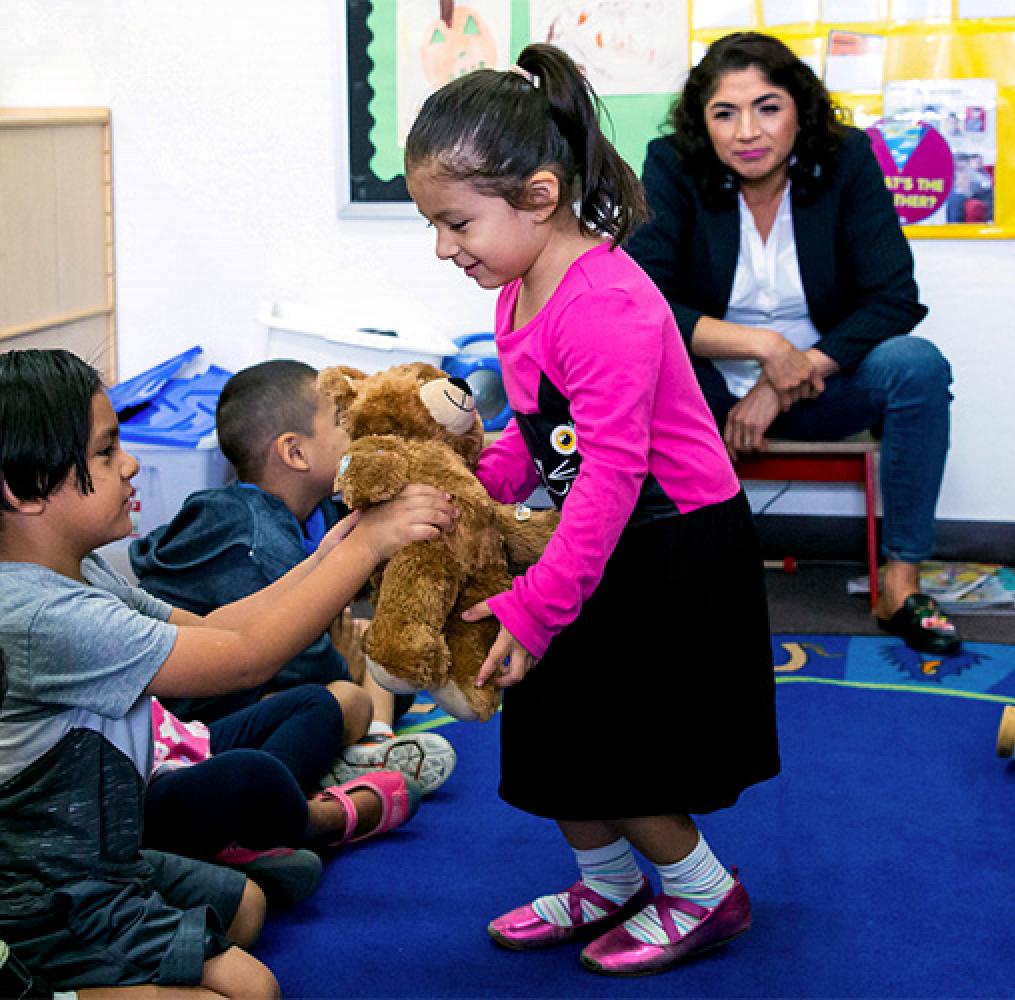 Elementary-aged girl hands a teddy bear to a classmate while a female teacher watches