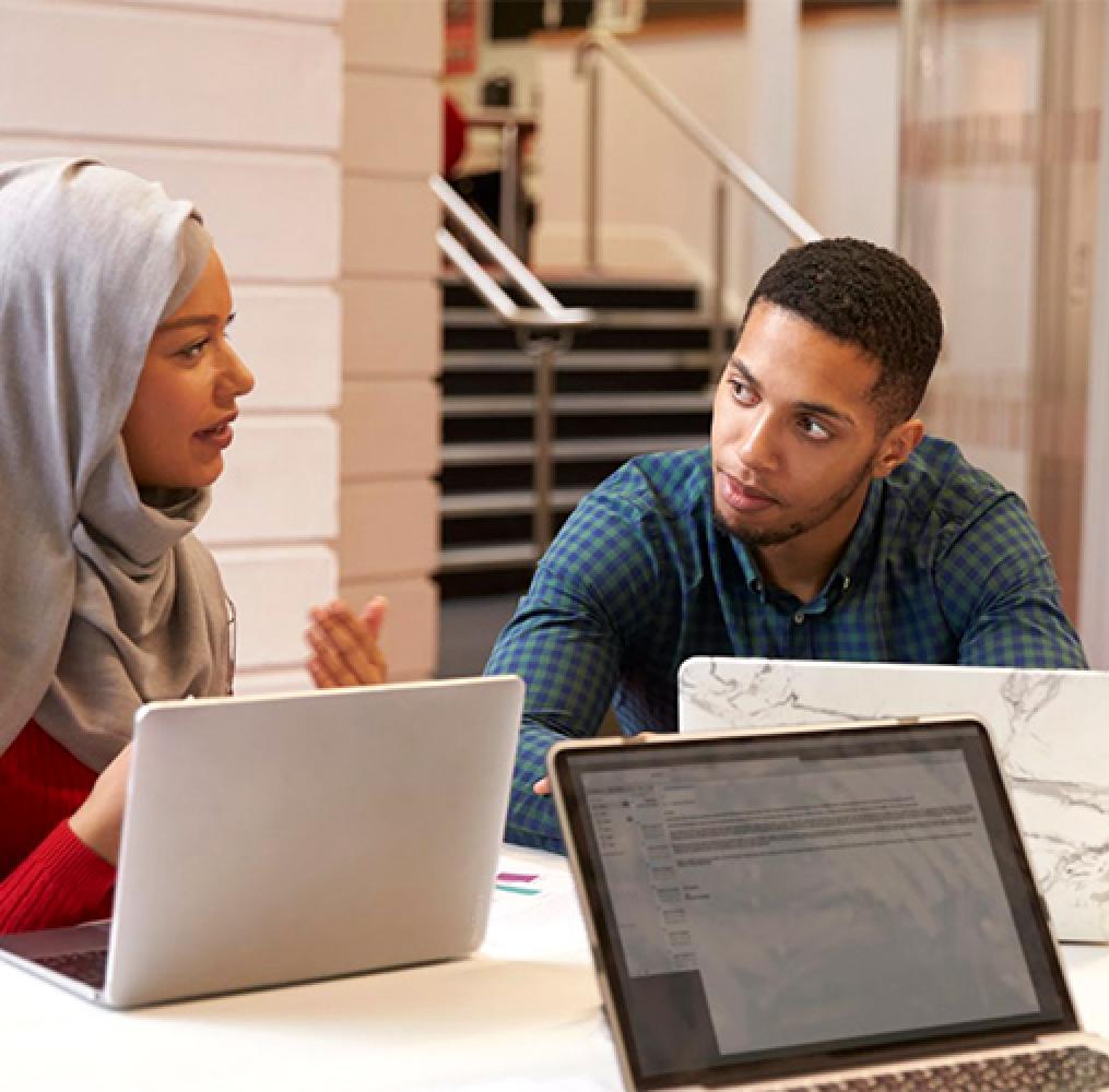 A woman wearing a headdress speaking with a man at a table with laptops