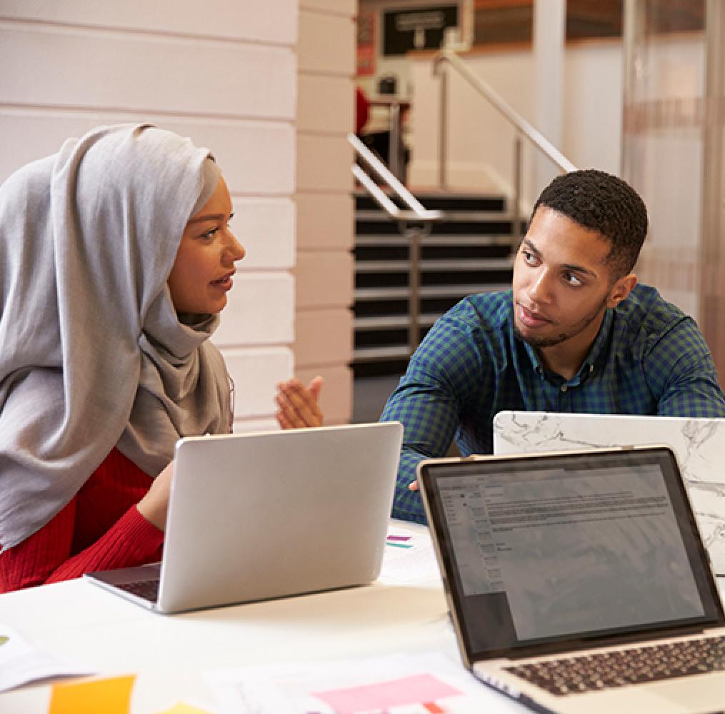 Woman wearing a head wrap is speaking with a male seated at a table with laptops