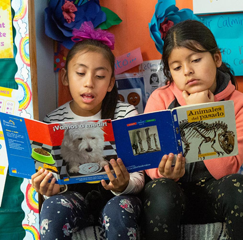 Two elementary girls reading Spanish-language books in a classroom.
