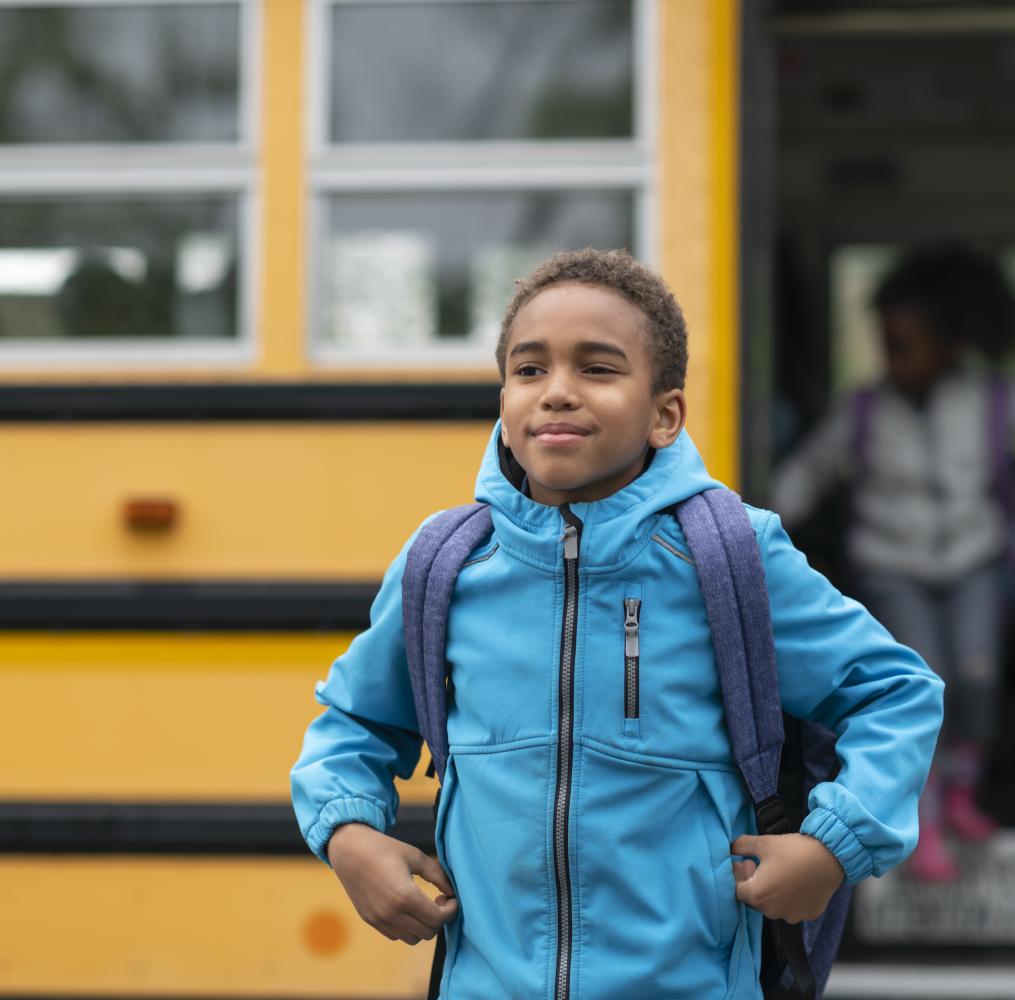 Elementary student smiling in front of a yellow school bus.