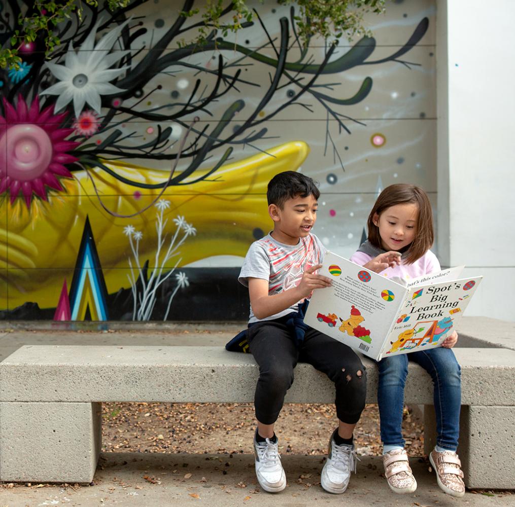 Two elementary students reading a book together while sitting on a bench.
