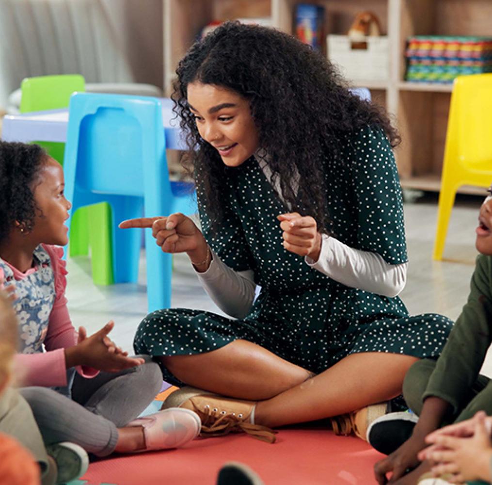 A teacher and elementary students seated on a rug in a classroom.