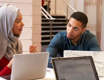 A woman wearing a headdress speaking with a man at a table with laptops