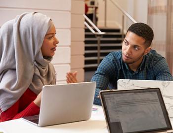 Woman wearing a head wrap is speaking with a male seated at a table with laptops