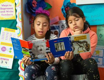 Two elementary girls reading Spanish-language books in a classroom.