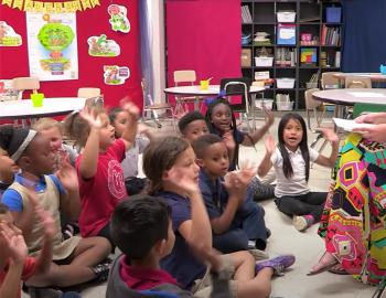 Elementary students raising their hands while seated on the ground in front of a teacher