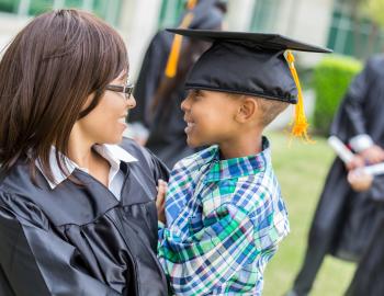 Woman in a graduation gown smiling with a child wearing a graduation cap.