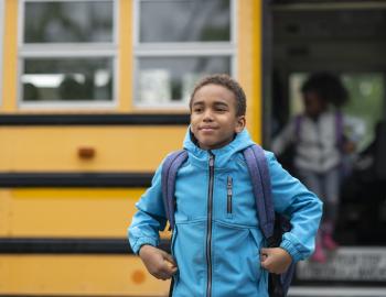 Elementary student smiling in front of a yellow school bus.