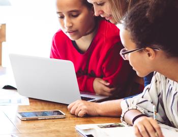 Teacher assisting two students with an assignment on a laptop.