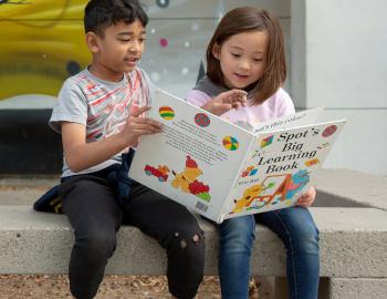Two elementary students reading a book together while sitting on a bench.